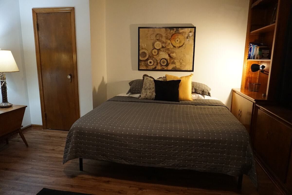 A bedroom with a bed, a wooden door, a wall-mounted artwork, a nightstand, and a bookshelf.