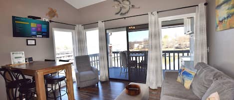 A living room with a wooden dining table, two chairs, a gray sofa, a ceiling fan, and a large window with curtains.