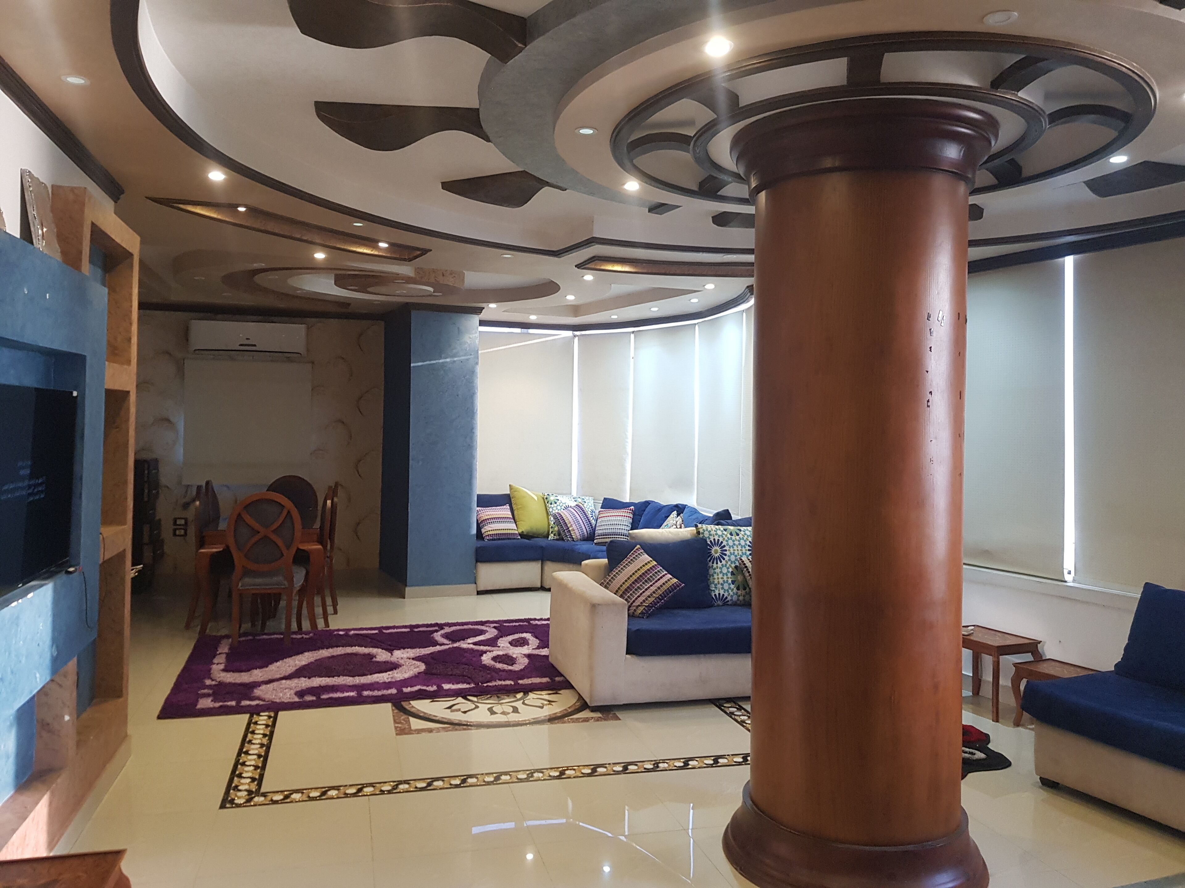 A modern living room with a large wooden column, a blue sofa with colorful cushions, a television, and a patterned rug.