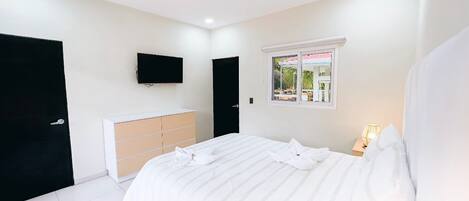 Un dormitorio con cama, televisor montado en la pared, ventana con vistas y mesita de noche con lámpara.