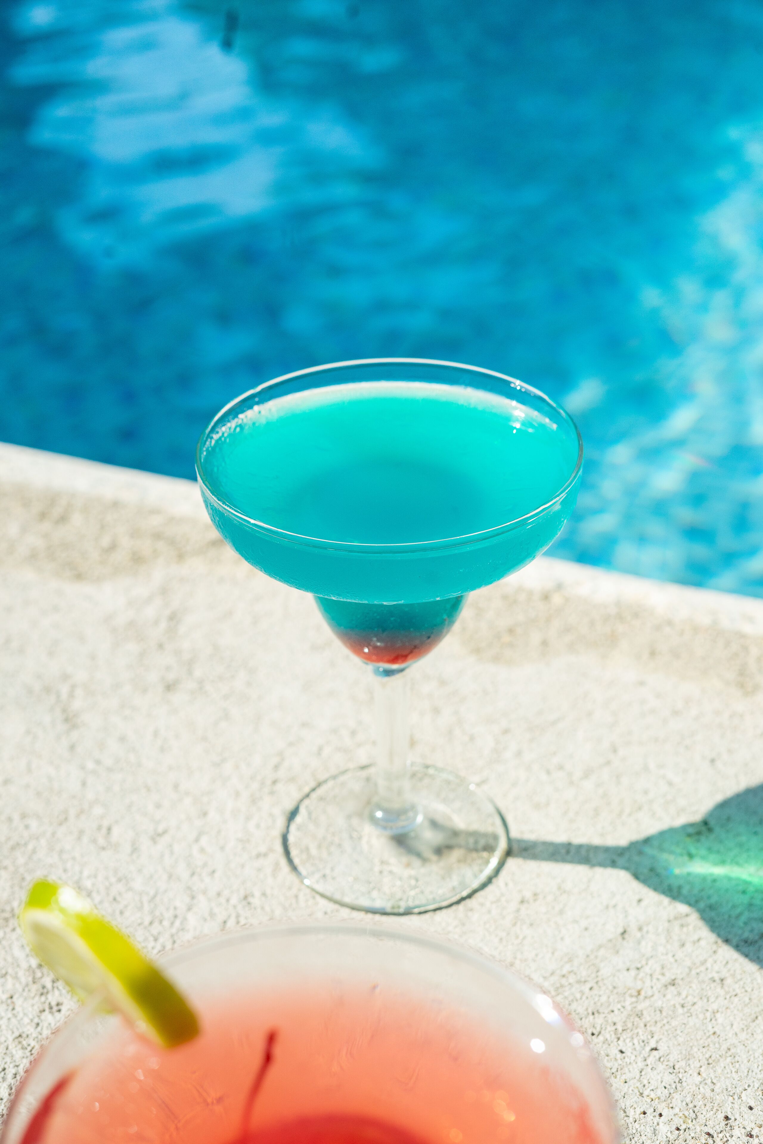 A turquoise cocktail in a martini glass with a lime garnish, placed on a concrete surface near a pool.