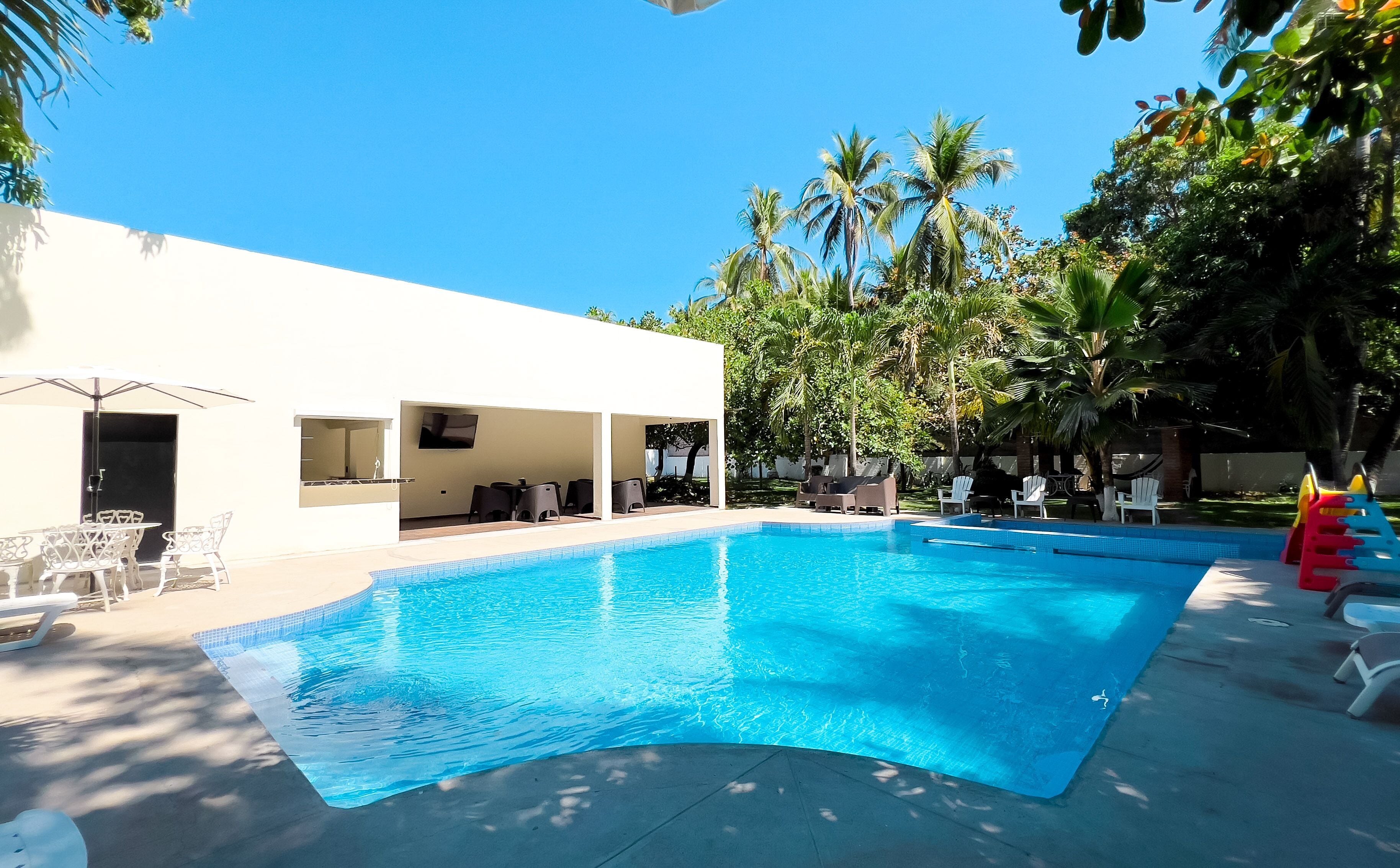 A swimming pool with lounge chairs and umbrellas, surrounded by palm trees and a white building with outdoor seating.