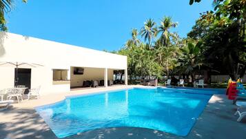A swimming pool with lounge chairs and umbrellas, surrounded by palm trees and a white building with outdoor seating.