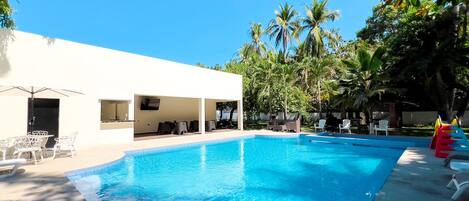 A swimming pool with lounge chairs and umbrellas, surrounded by palm trees and a white building with outdoor seating.