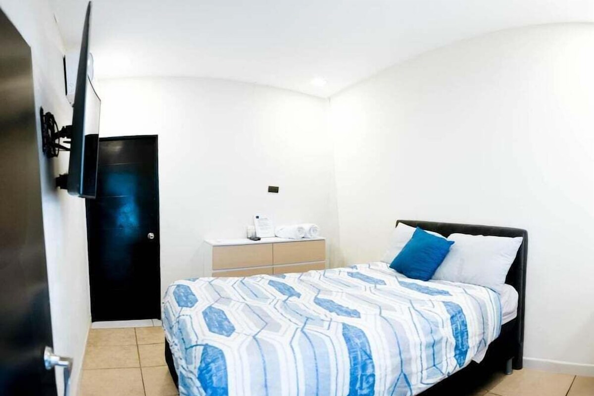 A bedroom with a bed, a bedside table, a TV mounted on the wall, and a door.