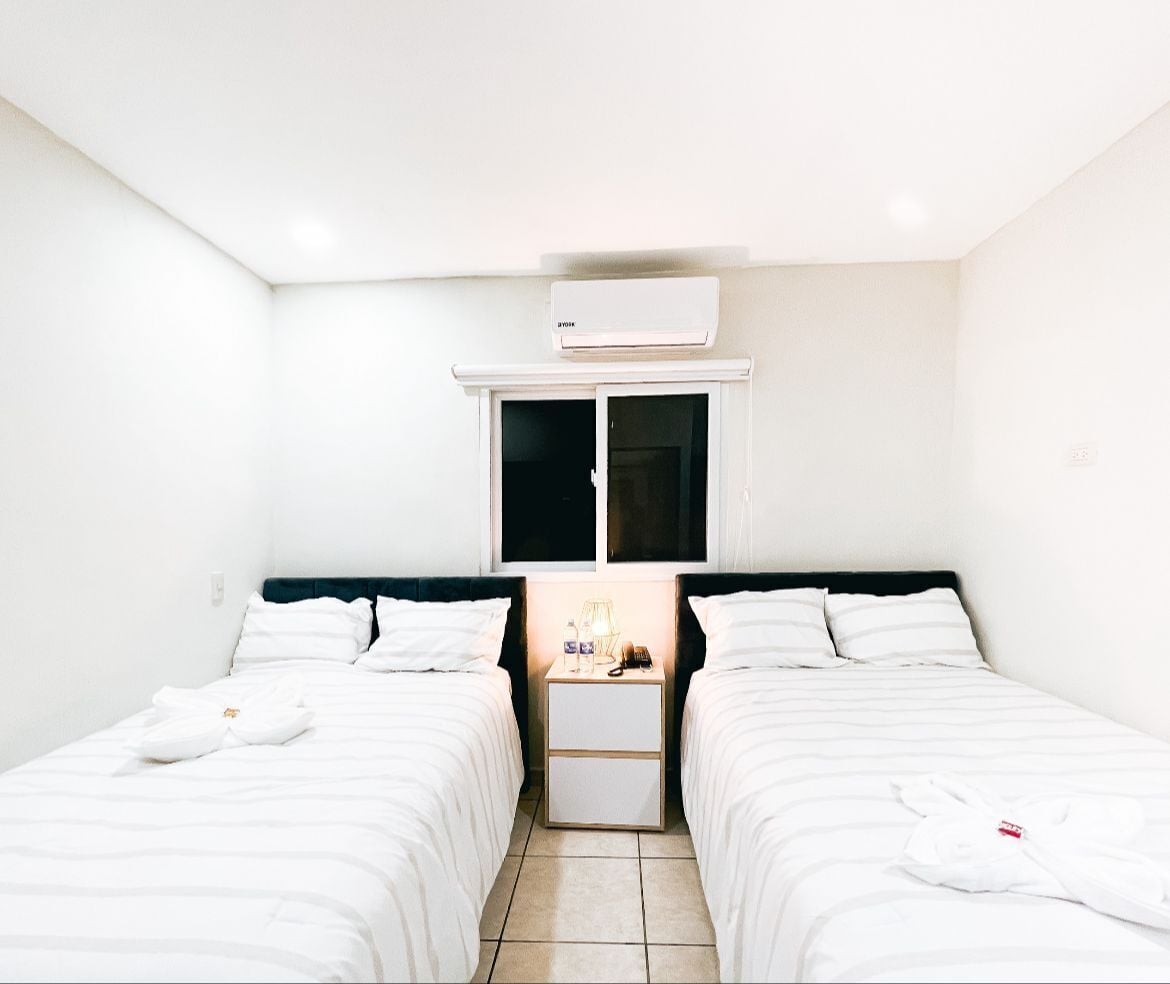 A hotel room with two beds, a nightstand with a lamp, and an air conditioning unit above the window.