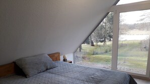 Une chambre avec un plafond en pente, un lit recouvert d’une courtepointe matelassée, une table de chevet avec une lampe, et une vue sur un espace verdoyant et des arbres, visible à travers une grande fenêtre.