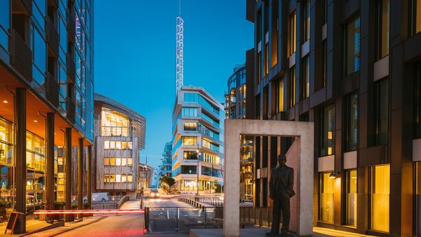 Una strada cittadina moderna al crepuscolo, con edifici contemporanei, una statua e un cielo sereno.