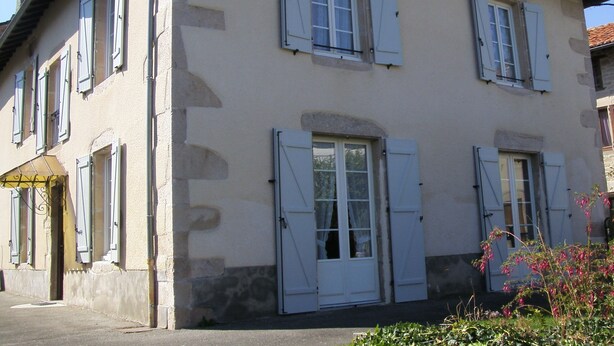 A two-story building with white shutters, a stone facade, and a small garden with pink flowers. - House with fenced garden in village (Latronquière)