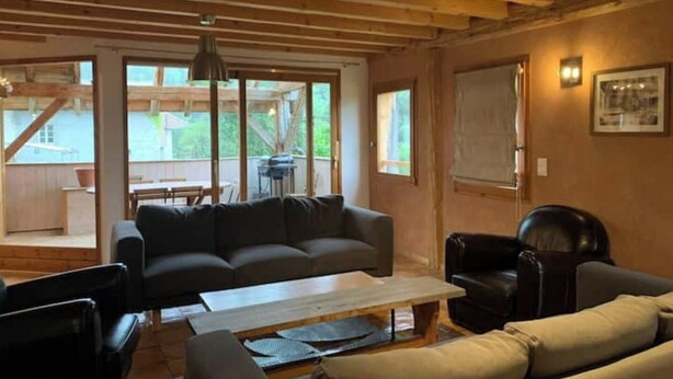 Ein gemütliches Wohnzimmer mit einer Couch, Sesseln und einem Couchtisch. Große Fenster bieten einen Blick ins Freie.