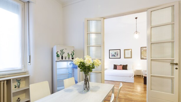 Un coin repas avec une table et des chaises blanches, un vase avec des fleurs, un réfrigérateur décoré d’aimants et, au fond, un canapé blanc.