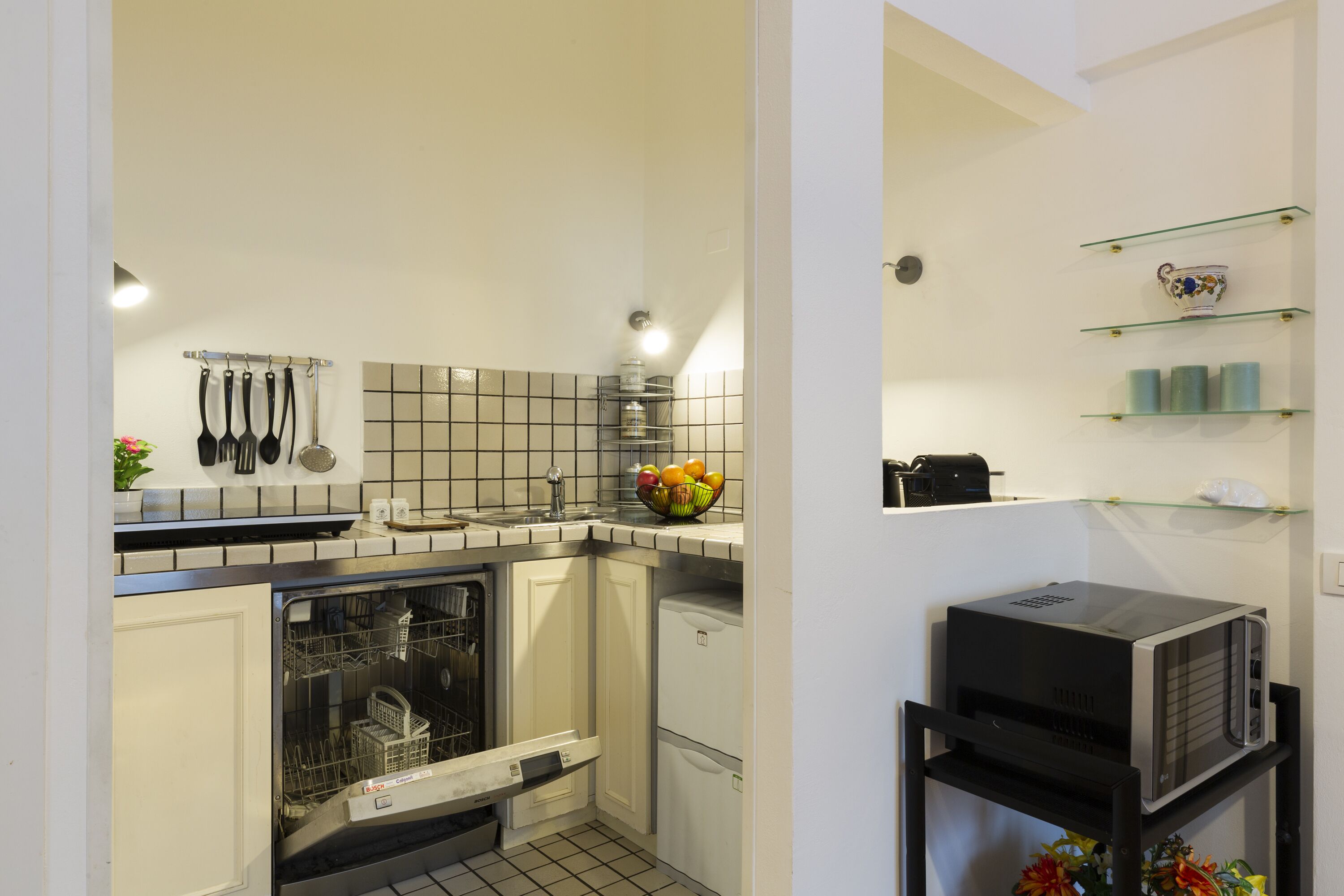 A compact kitchen with a dishwasher, a small refrigerator, and a microwave on a black countertop. There are hanging utensils and a fruit bowl on the counter.