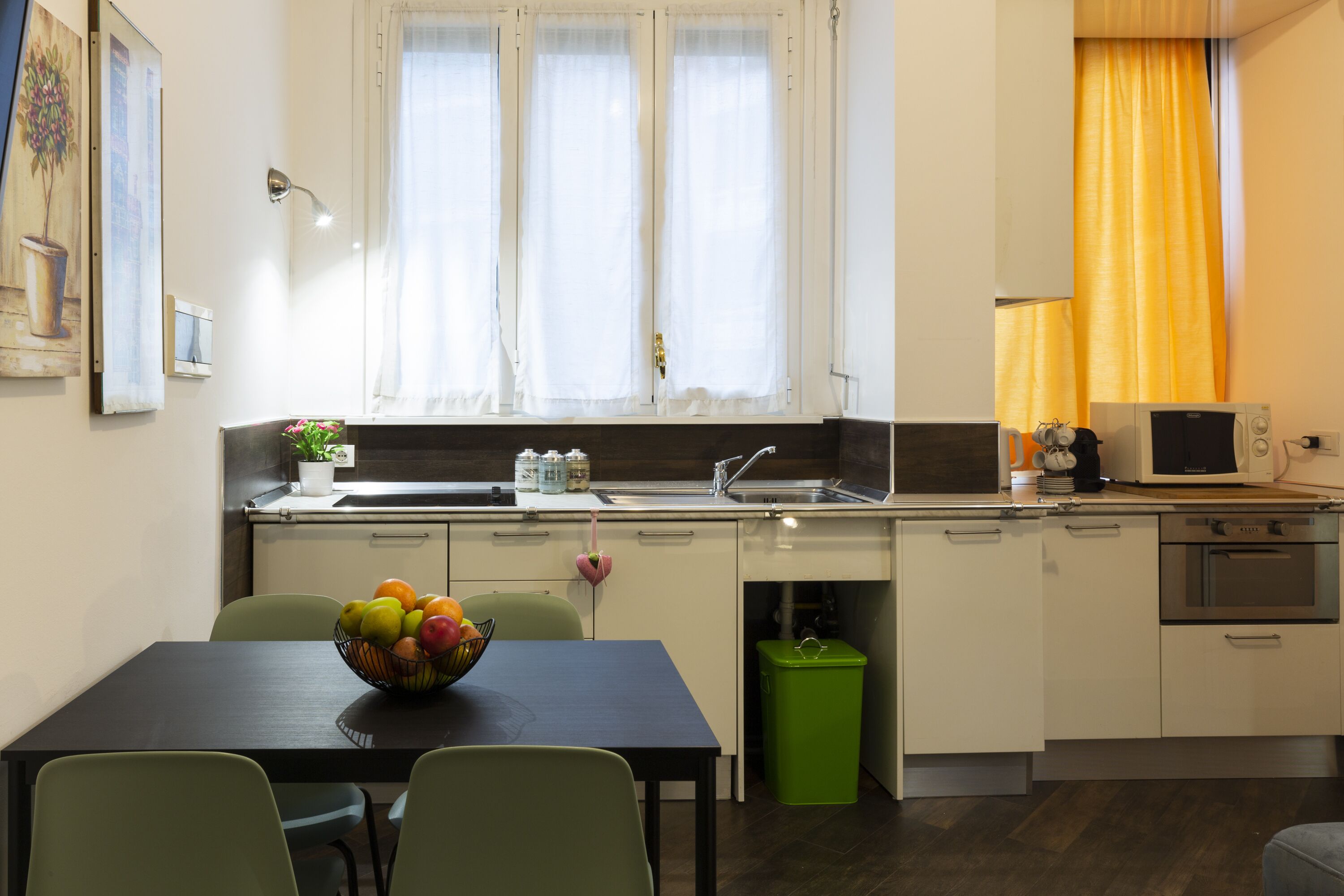 A compact kitchen with a dining table, chairs, a fruit bowl, a sink, a microwave, and a green trash bin.