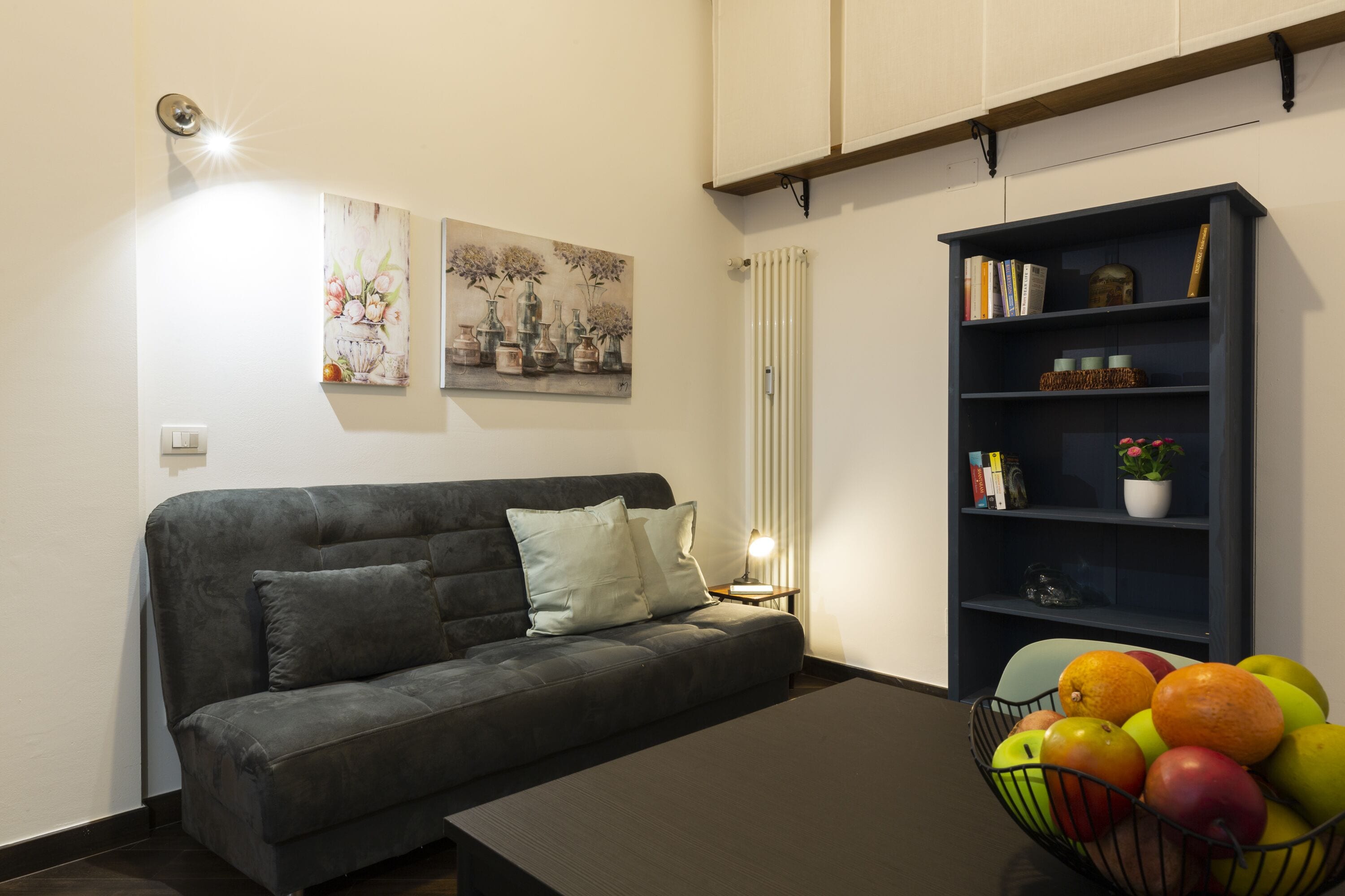 A living room with a dark grey sofa, a coffee table, a bookshelf, and a fruit bowl.