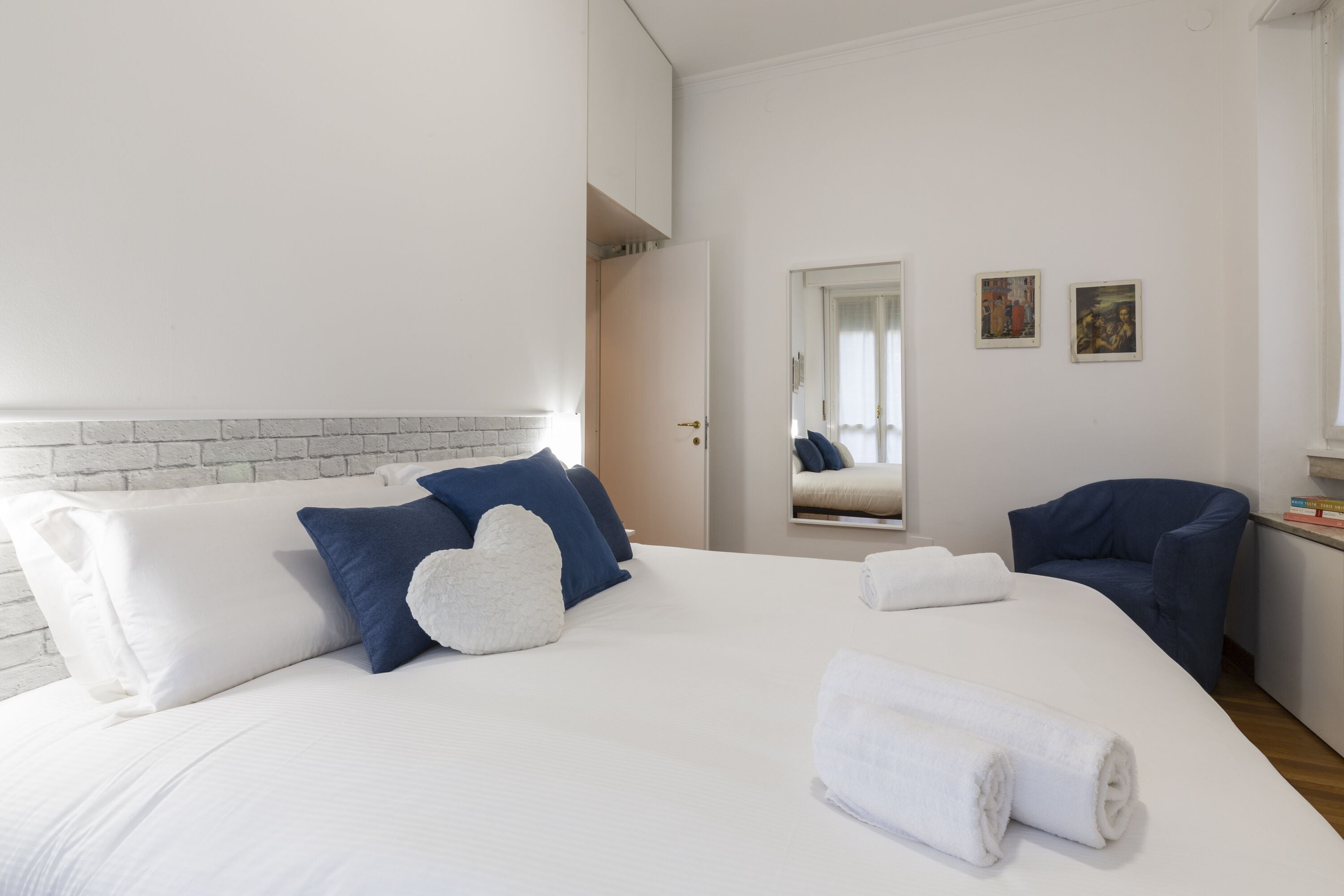 Camera d'albergo con un letto grande, biancheria azzurra e bianca, una poltrona blu e un tavolino con dei libri.
