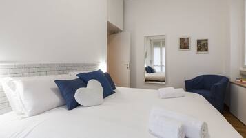 Camera d'albergo con un letto grande, biancheria azzurra e bianca, una poltrona blu e un tavolino con dei libri.