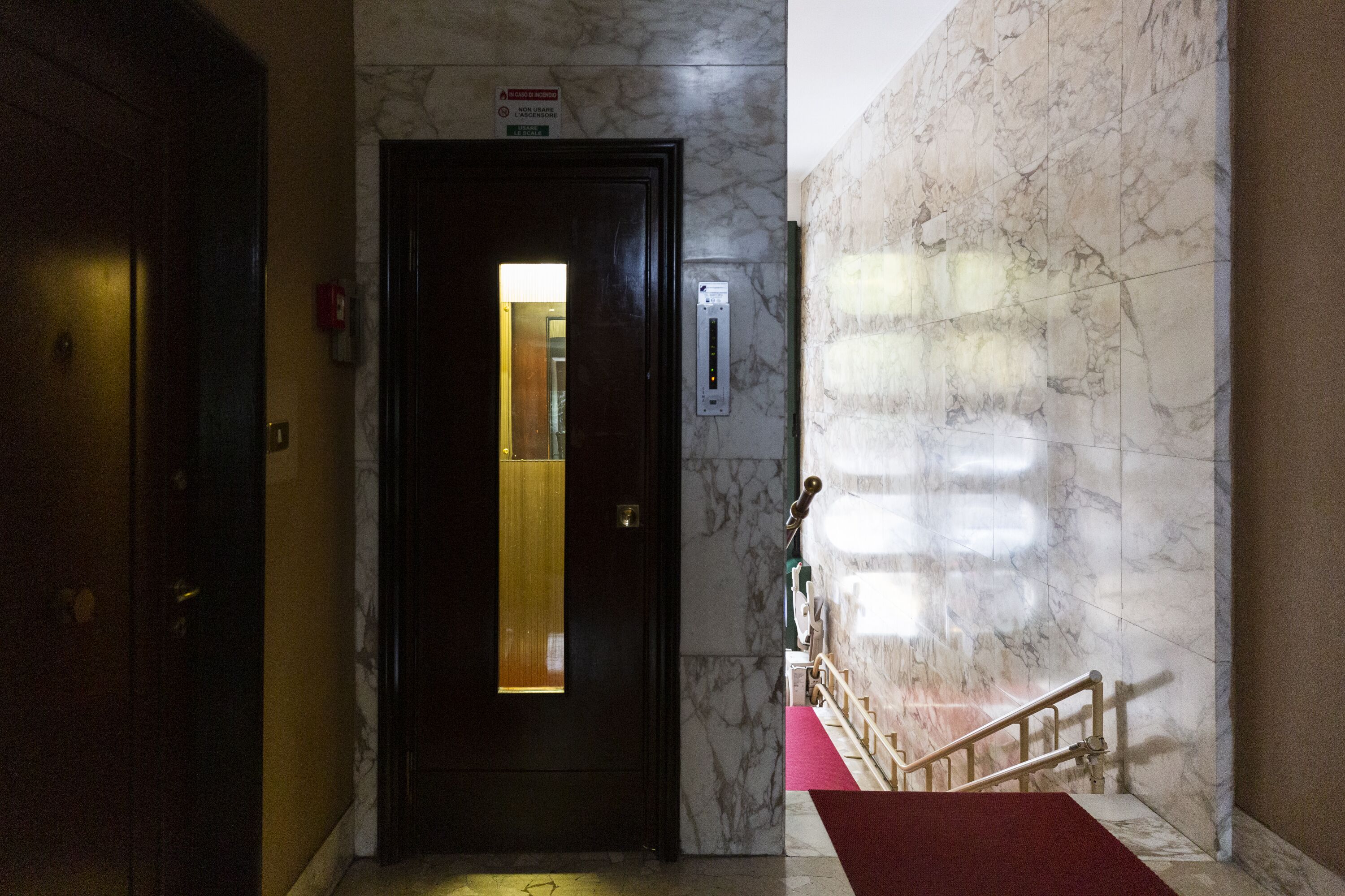 A hotel lobby with a marble wall, a red carpet, and a door with a lighted panel.