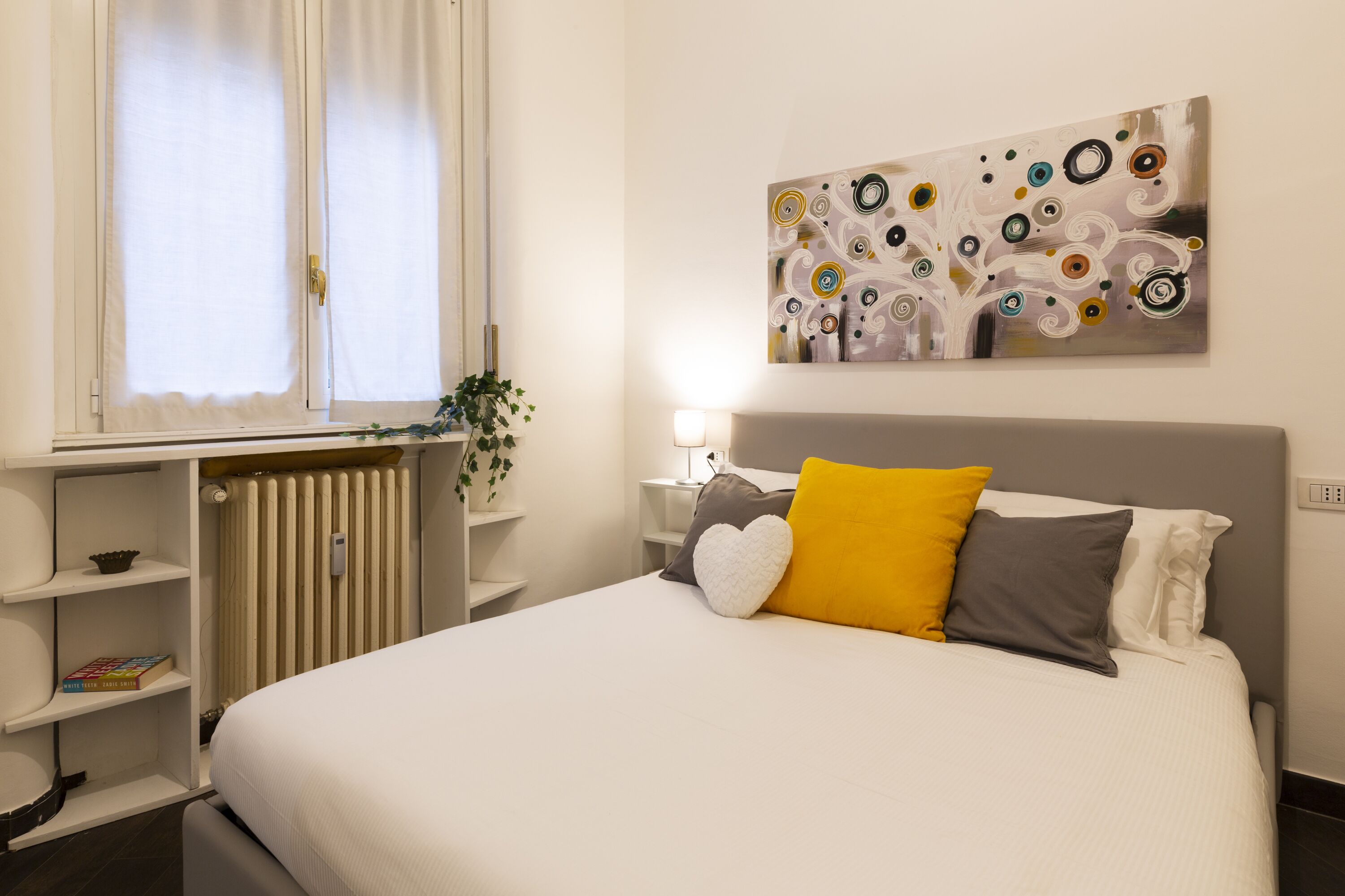 A neatly arranged bedroom with a bed, bedside tables, a radiator, and a window with curtains.