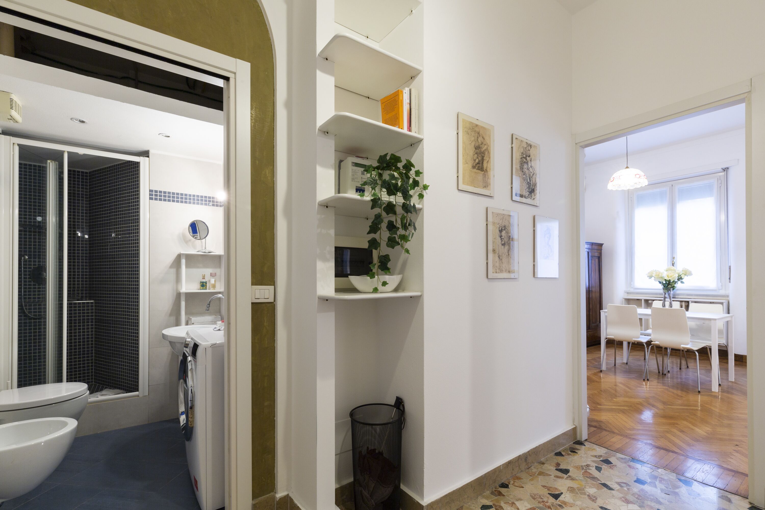 A modern bathroom with a shower, toilet, and sink. There is a hallway leading to a dining area with a table and chairs.