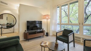 Ein modernes Wohnzimmer mit Flachbildfernseher, einer Couch, einem Sessel, einem Couchtisch und großen Fenstern mit Blick auf Bäume.