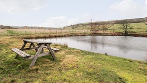 A picnic table by a calm pond with grassy banks and distant buildings. - Rivendell (Holmfirth)
