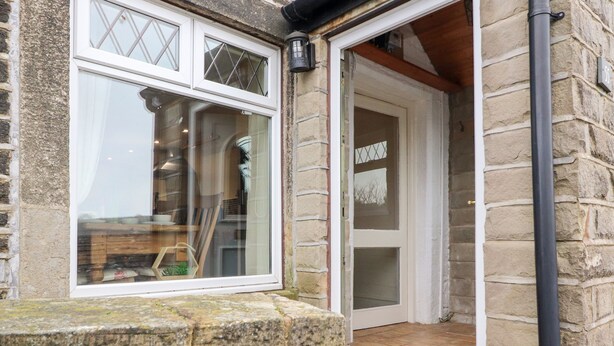 A stone building with a white-framed window and a door leading to a tiled entrance. - Rivendell (Holmfirth)