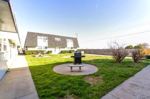 A grassy courtyard with a bench, a trash can, and a building in the background.
