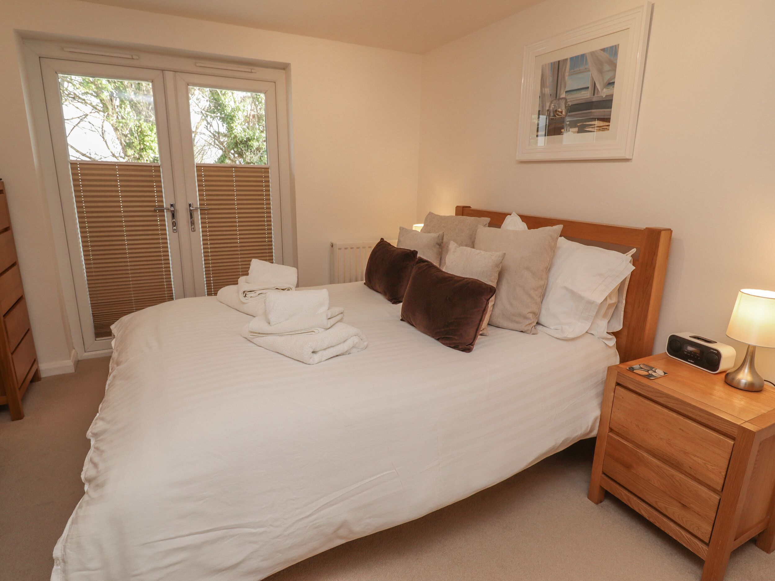 A neatly made bed with white linens and brown pillows, a wooden nightstand with a radio, and a door with blinds.
