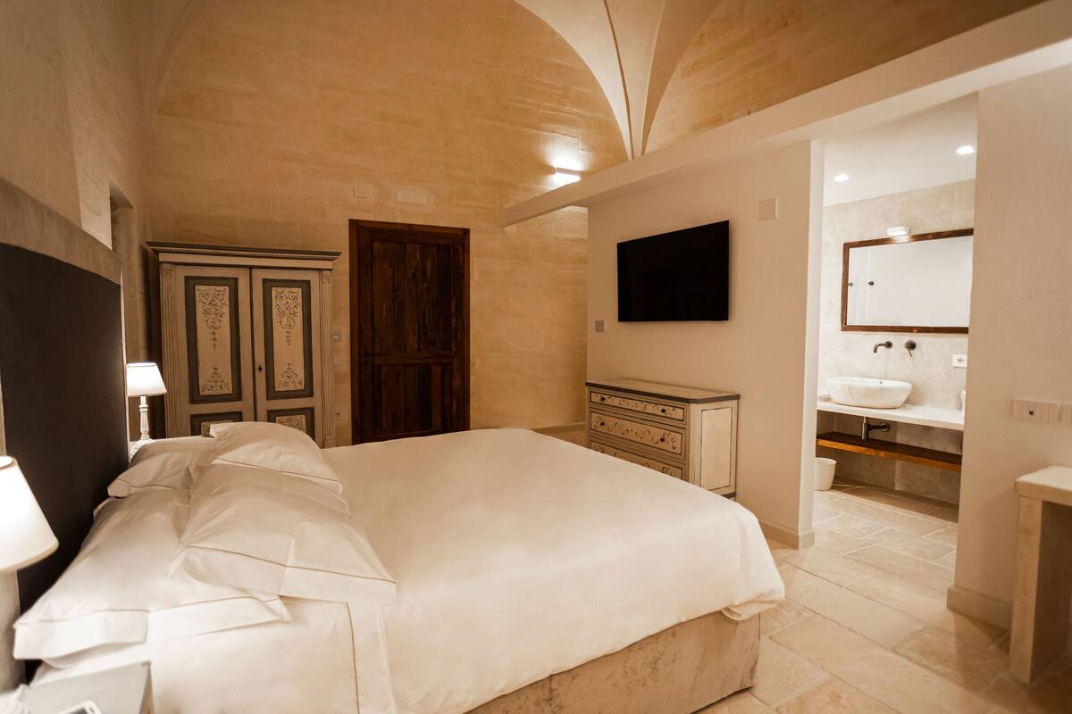 Un dormitorio amplio con una cama grande, un armario de madera, un televisor instalado en la pared y un baño con lavamanos y espejo.