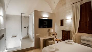 Quarto de hotel com cama, escrivaninha, televisão, chuveiro e guarda-roupa.