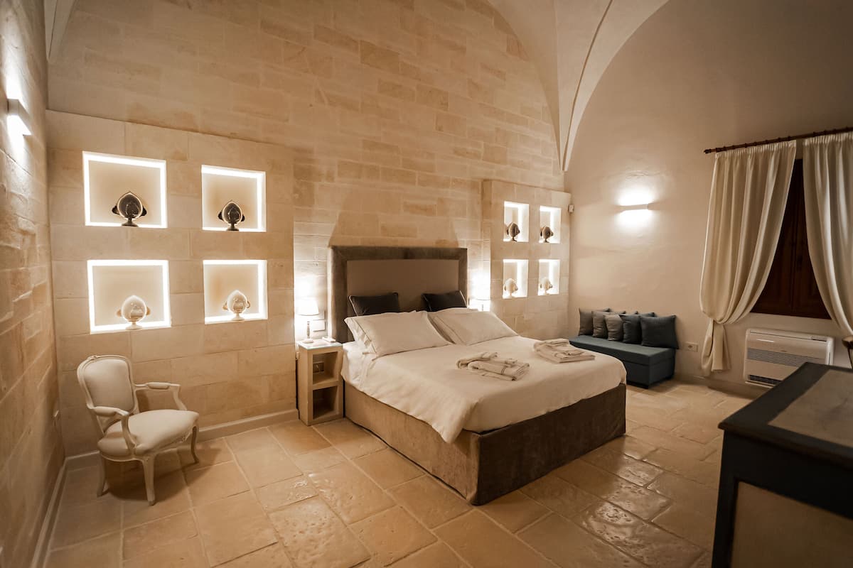 Un dormitorio con pared de piedra, una cama con almohadas, una silla y mesitas de noche con lámparas.
