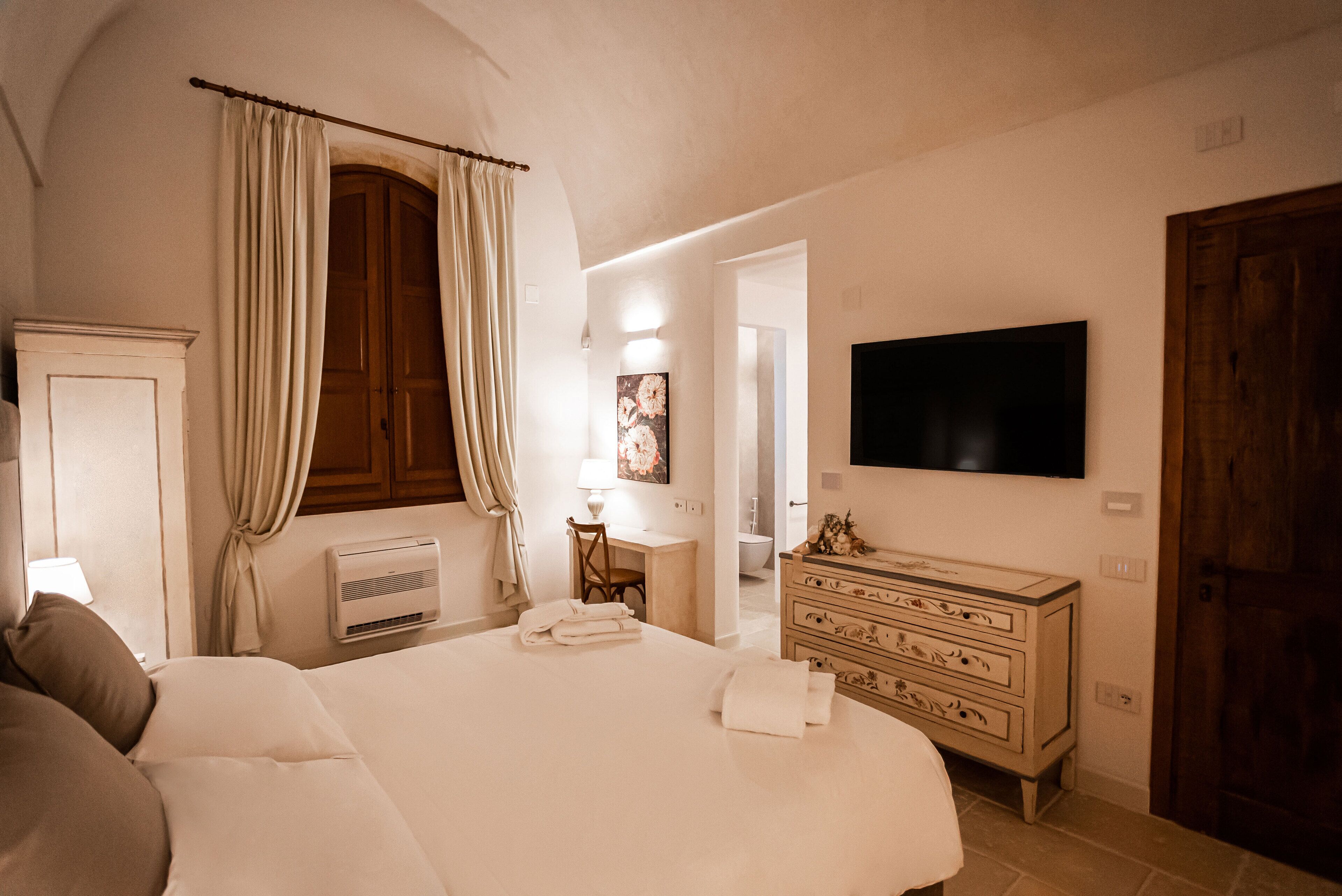 A bedroom with a large bed, a dresser, a television, and a bathroom visible through an open door.