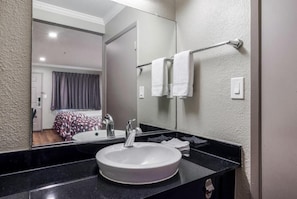 Une salle de bain avec un lavabo rond, un miroir, un porte-serviettes et un lit que l’on aperçoit à travers le miroir.