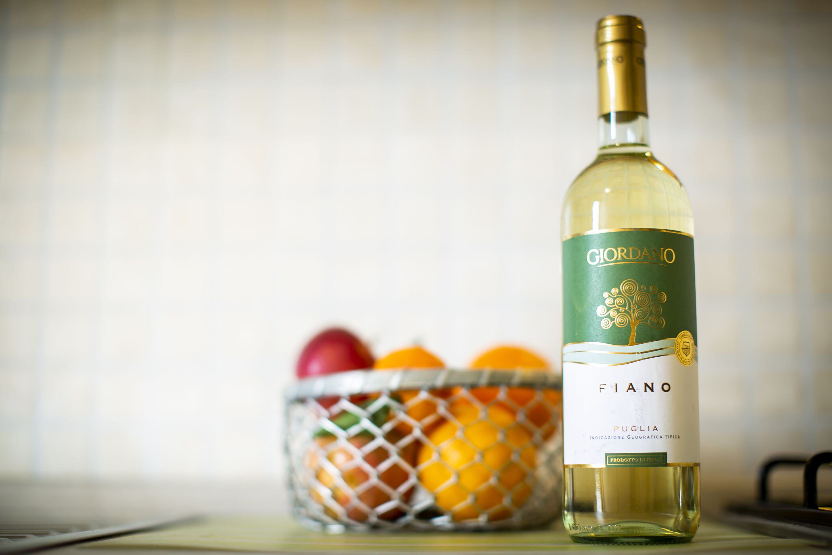 Una botella de vino con etiqueta que dice "GIORDANO FIANO PUGLIA" junto a una cesta de frutas.