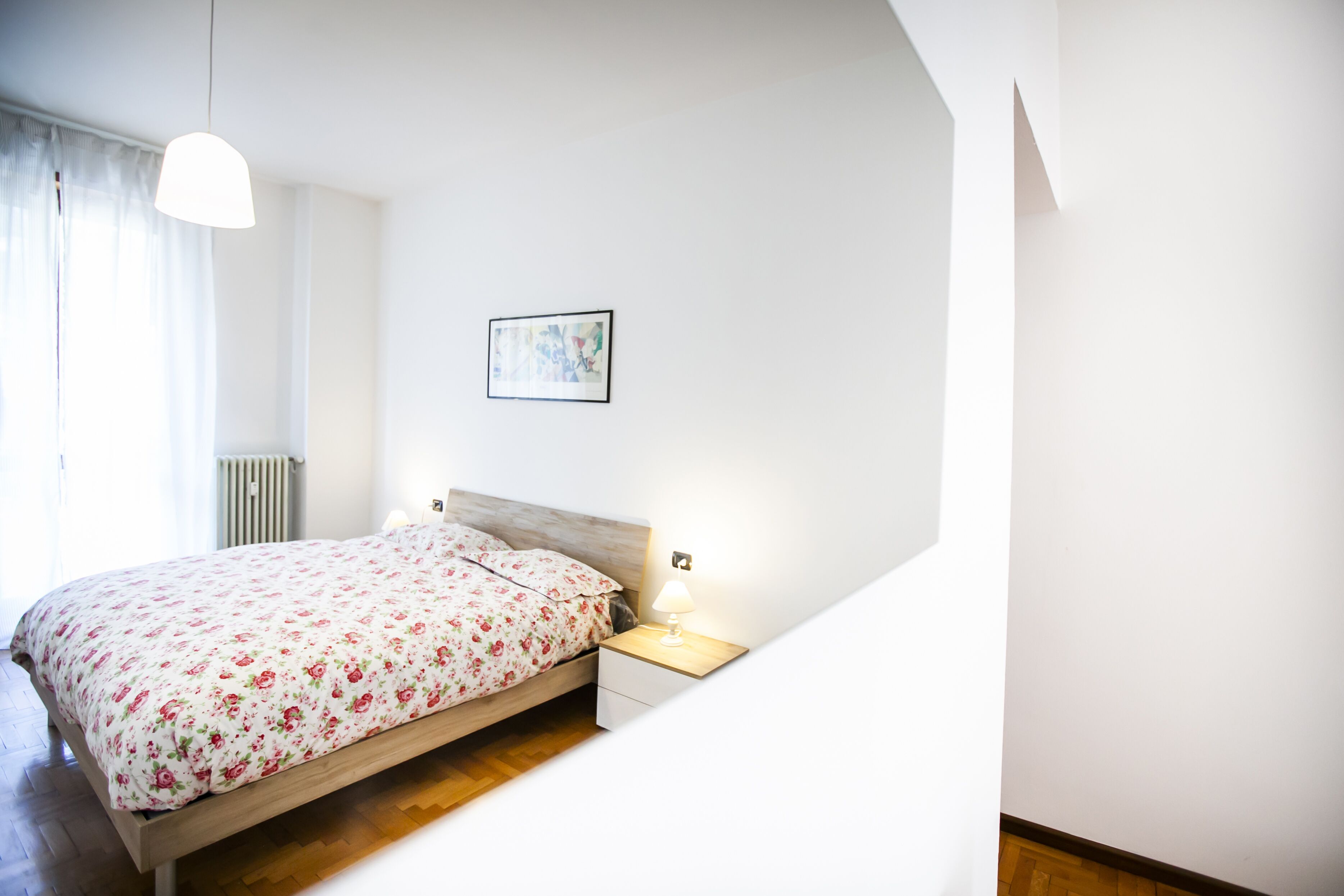 A bedroom with a wooden bed, a bedside table with a lamp, a radiator, and a window with curtains.