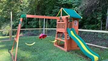A wooden play structure with a slide and swings, surrounded by a fence and greenery.