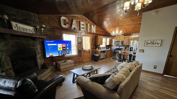 A cabin interior with a stone fireplace, a television, a sofa, and a kitchen area. - Log Cabin Minutes to Dollywood all Pigeon Forge (Sevier County)