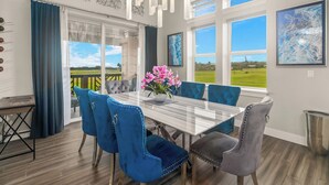 Un coin repas avec une table en verre, des chaises bleues et grises, un vase rempli de fleurs roses et de grandes fenêtres donnant sur un paysage verdoyant.