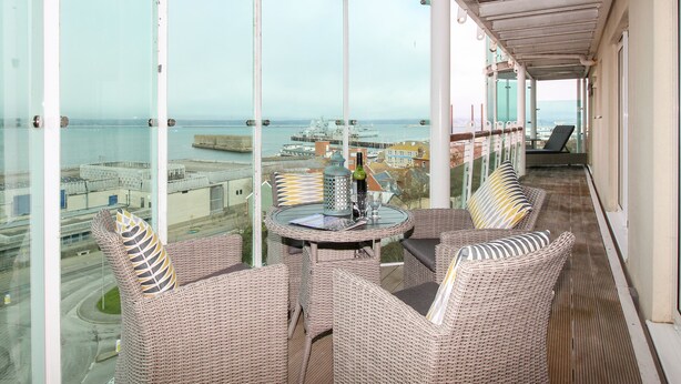 A balcony with wicker furniture, a glass table, and a view of the sea and buildings. - Bear's Apartment (Portland)