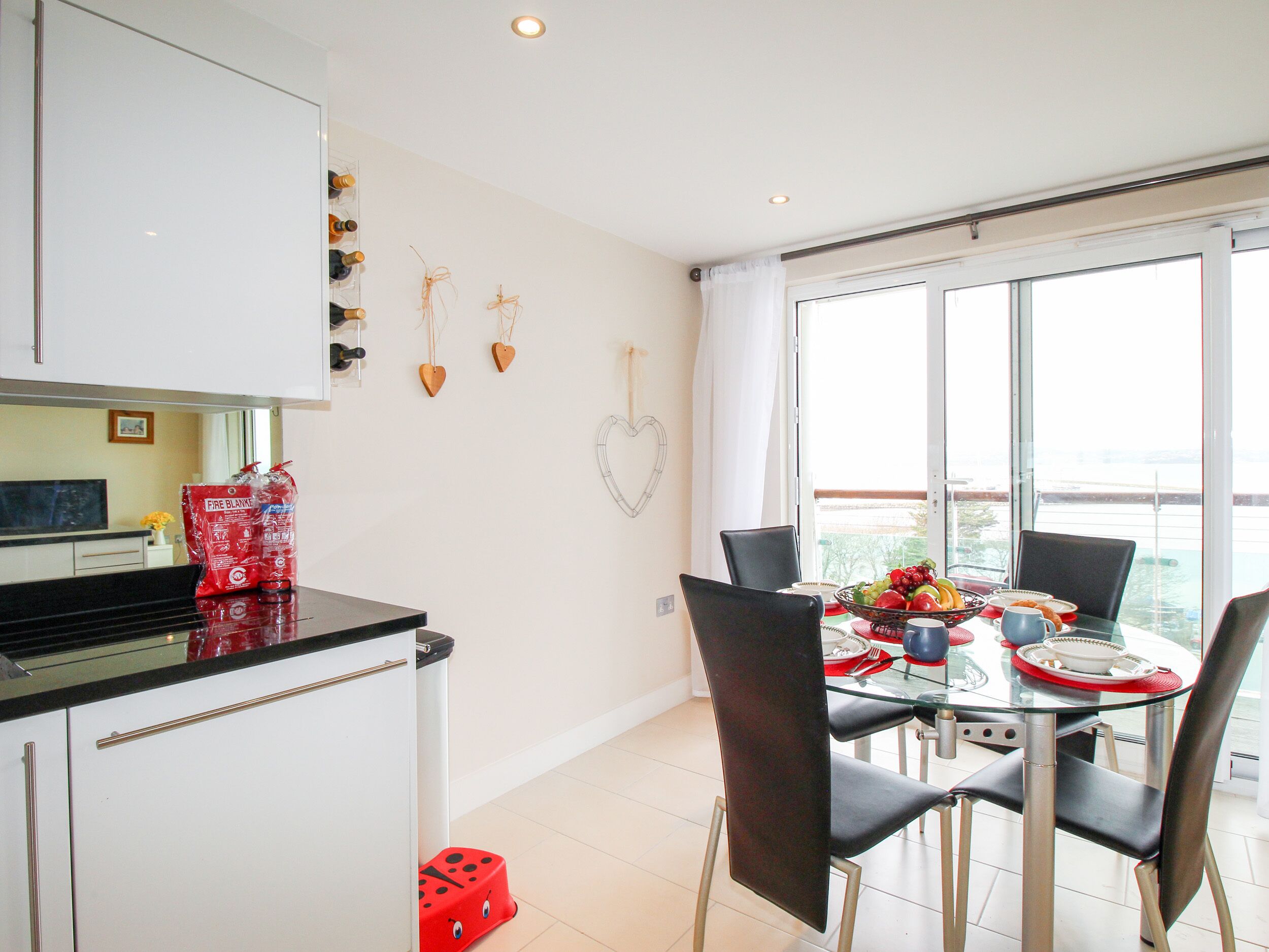 A modern kitchen with white cabinets, a glass dining table, and chairs. There is a window with a view of the sea.