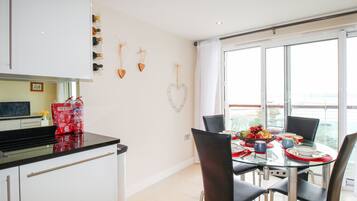 A modern kitchen with white cabinets, a glass dining table, and chairs. There is a window with a view of the sea.