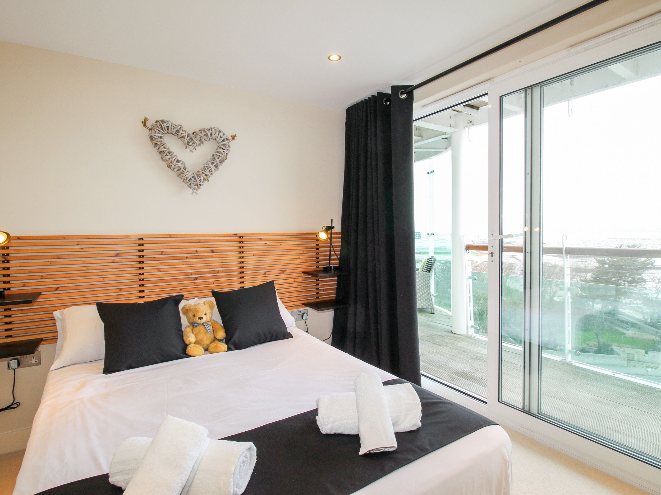 A hotel room with a large bed, a teddy bear on the pillows, a heart-shaped decoration on the wall, and a balcony with a view.