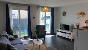 Ett modernt vardagsrum med en soffa, en fåtölj, ett litet bord, en tv på ett vitt skåp och en balkong med blå gardiner.