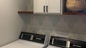 A laundry area with two front-loading washing machines, a white cabinet above, and a potted plant on a shelf. - Luxury Living at The Harrison (Le Mars)