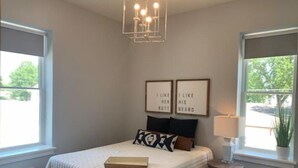 A bedroom with a bed, a bedside table with a lamp, a window with a view, and two framed pictures on the wall. - Luxury Living at The Harrison (Le Mars)