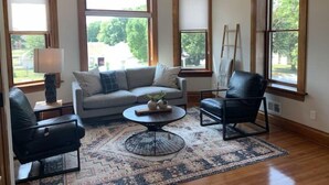 A spacious living room with a large window, a sofa, a chair, a coffee table, and a chandelier. - Luxury Living at The Harrison (Le Mars)