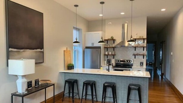 A modern kitchen with a central island, bar stools, pendant lighting, and a view into the adjacent living area. - Luxury Living at The Harrison (Le Mars)
