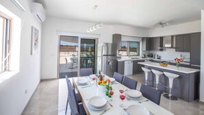 A modern kitchen with a central island, a dining area with a table set for four, and a view of the outdoors through the sliding doors. - Villa Demeter GA98 - Six Bedroom Villa, Sleeps 12 (Paralimni)