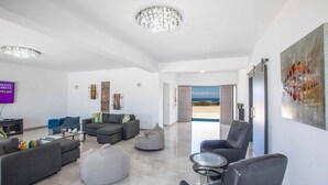 A spacious living room with a large sofa, a coffee table, and a bean bag. There is a television on the left and a door leading to a balcony. - Villa Demeter GA98 - Six Bedroom Villa, Sleeps 12 (Paralimni)