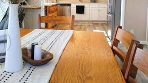 Un coin repas avec une table en bois, des chaises, un vase contenant des plantes séchées et, au fond, une cuisine.